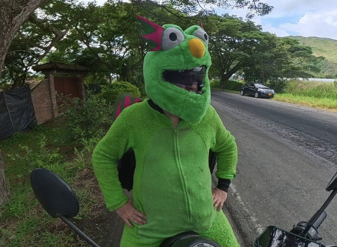 Fluffy standing hands on hips beside his motorcycle on a tree-lined rural road in Colombia